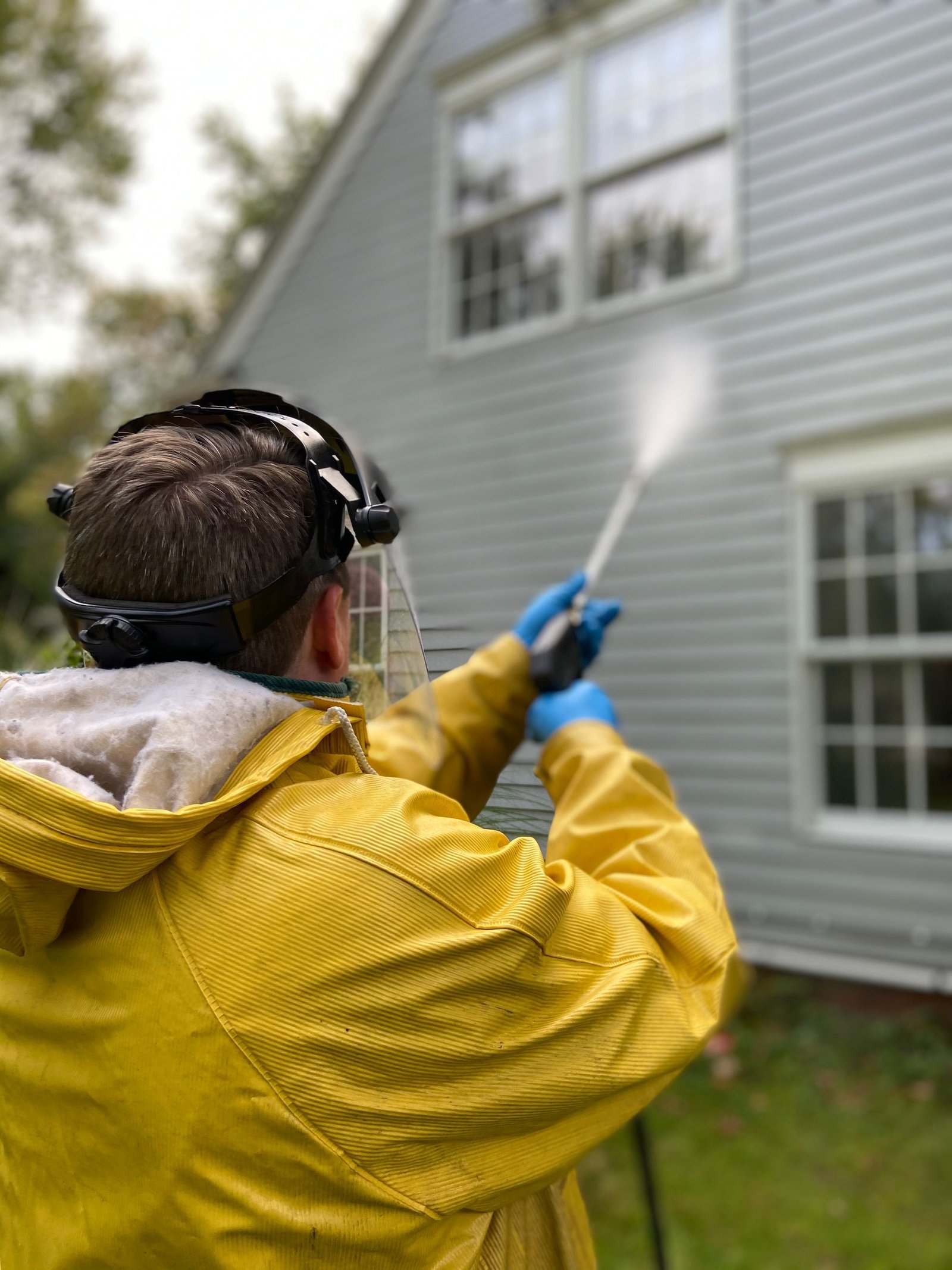 Een persoon met gele jas en blauwe handschoenen spuit de gevel van een huis schoon met hogedruk